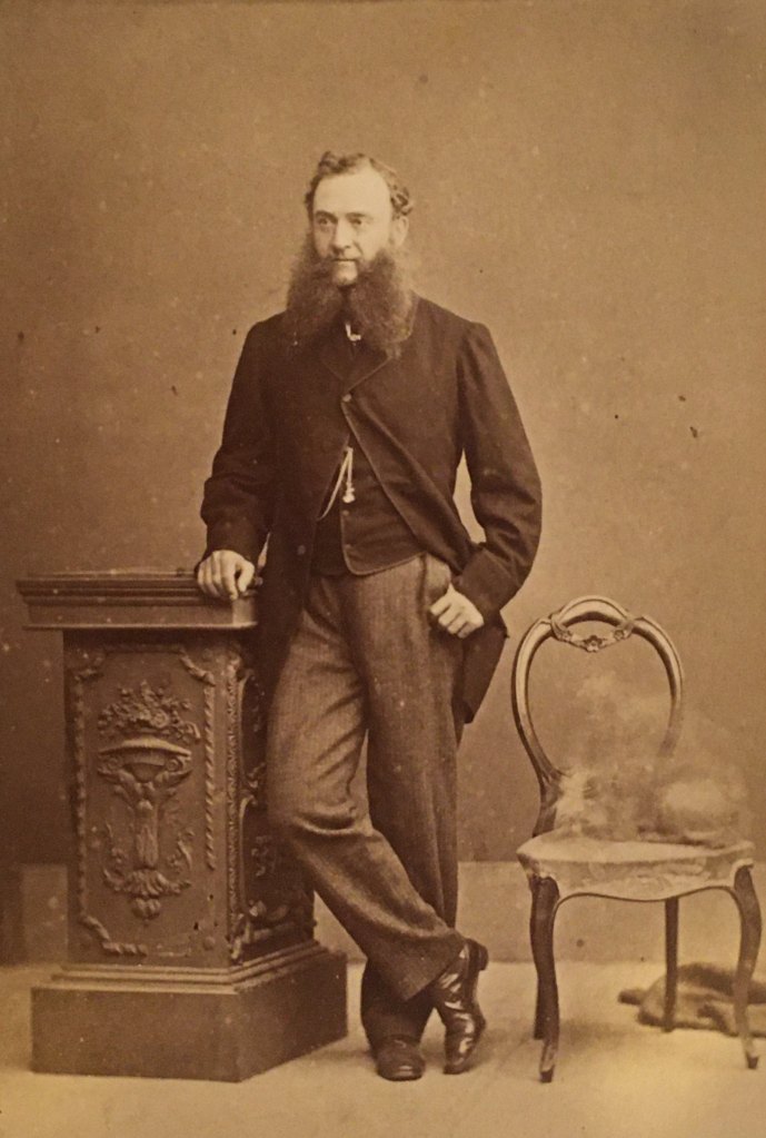 A cabinet card of a man with a forked beard, standing for a photograph. A blur on the chair next to him suggests that an animal is present.